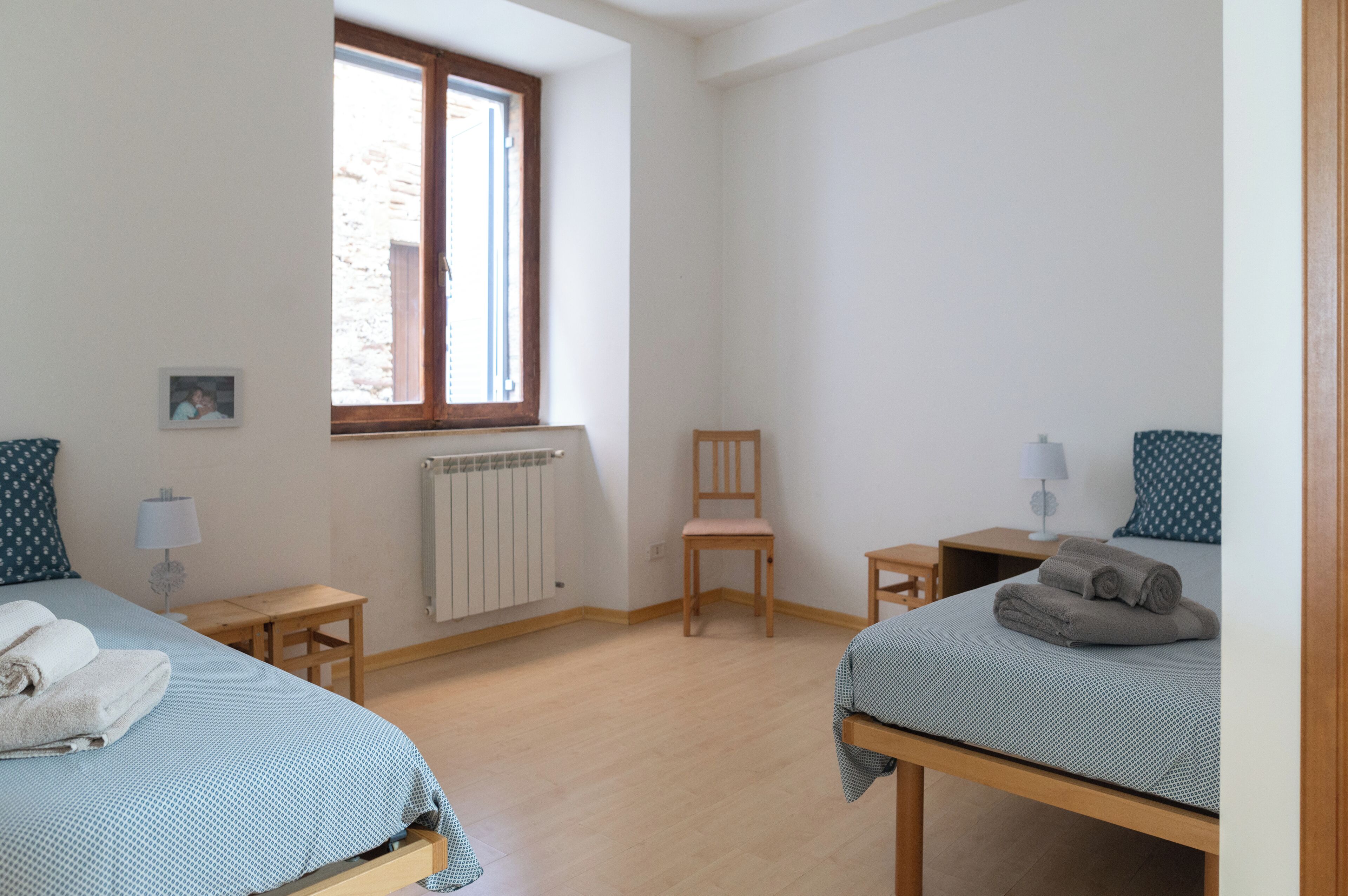 Quarto com duas camas, uma janela, uma cadeira, uma mesa e um radiador.