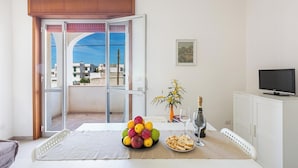 A room with a balcony, a table with fruit, a bottle of champagne, and a TV on a cabinet. - 3872 Residence Kalura - Bilo 6 by Barbarhouse (Porto Cesareo)