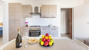 A modern kitchen with light wood cabinets, a gas stove, and a sink. A table with a bottle of wine, glasses, and a fruit bowl is in the foreground. - 3872 Residence Kalura - Bilo 6 by Barbarhouse (Porto Cesareo)