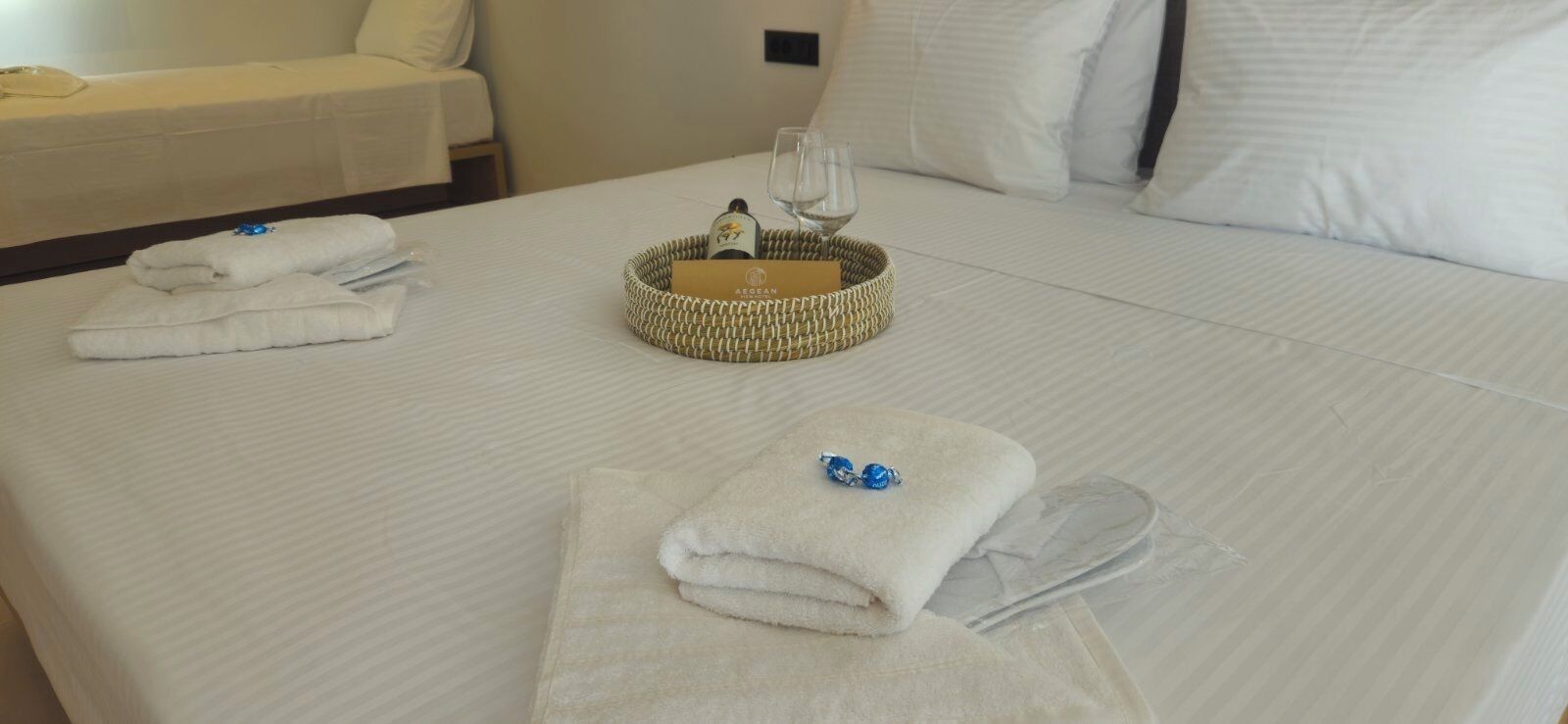 A neatly made bed with white linens, a woven basket with a bottle and glasses, and a folded towel with a blue item on it.