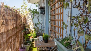 Een klein terras buiten met een houten tafel en stoelen, omgeven door potplanten en een bamboomscherm.