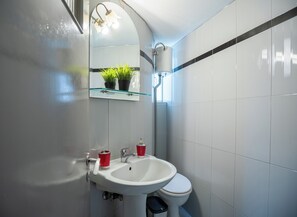 A bathroom with a white sink, a toilet, and a glass shelf with plants. - Acropolis Rooftop View! 10 Minutes From Everywhere (Athina)