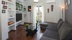 Een woonkamer met een bank, televisie, planken en een glazen salontafel.