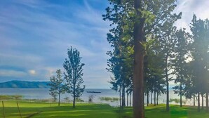 A grassy area with trees and a body of water in the background. - Eben Lake Kivu Cottages and Villas (Kagano)