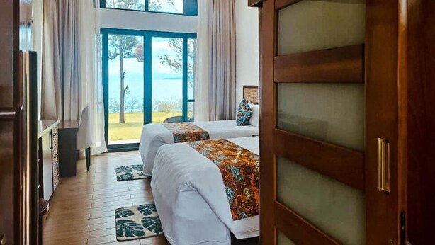 A hotel room with two beds, a wooden wardrobe, a desk, and a view of the sea through a glass door. - Eben Lake Kivu Cottages and Villas (Kagano)