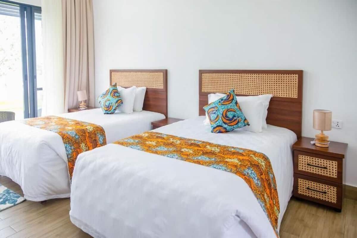 Une chambre d’hôtel avec deux lits, chacun doté d’une tête de lit en bois et de linge de lit blanc. Chaque lit est agrémenté d’une table de chevet avec une lampe et d’un coussin décoratif.