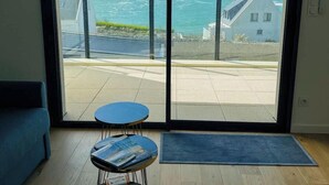 A room with a large sliding glass door leading to a balcony with a view of the sea and buildings.