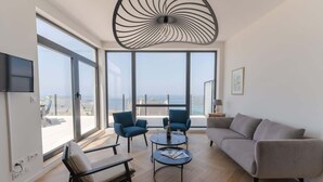 Una sala de estar moderna con ventanas amplias, un sofá gris, dos sillones azules, una mesa de centro redonda y una lámpara de techo original.