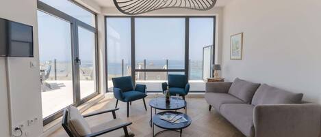 A modern living room with large windows, a grey sofa, two blue armchairs, a round coffee table, and a unique ceiling light fixture.