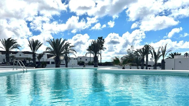 A clear blue swimming pool surrounded by palm trees and white buildings under a partly cloudy sky. - Fkk-apartment in Villa Blanca, 4 mit Schwimmbad und Zwei Schlafzimmer (Charco del Palo)