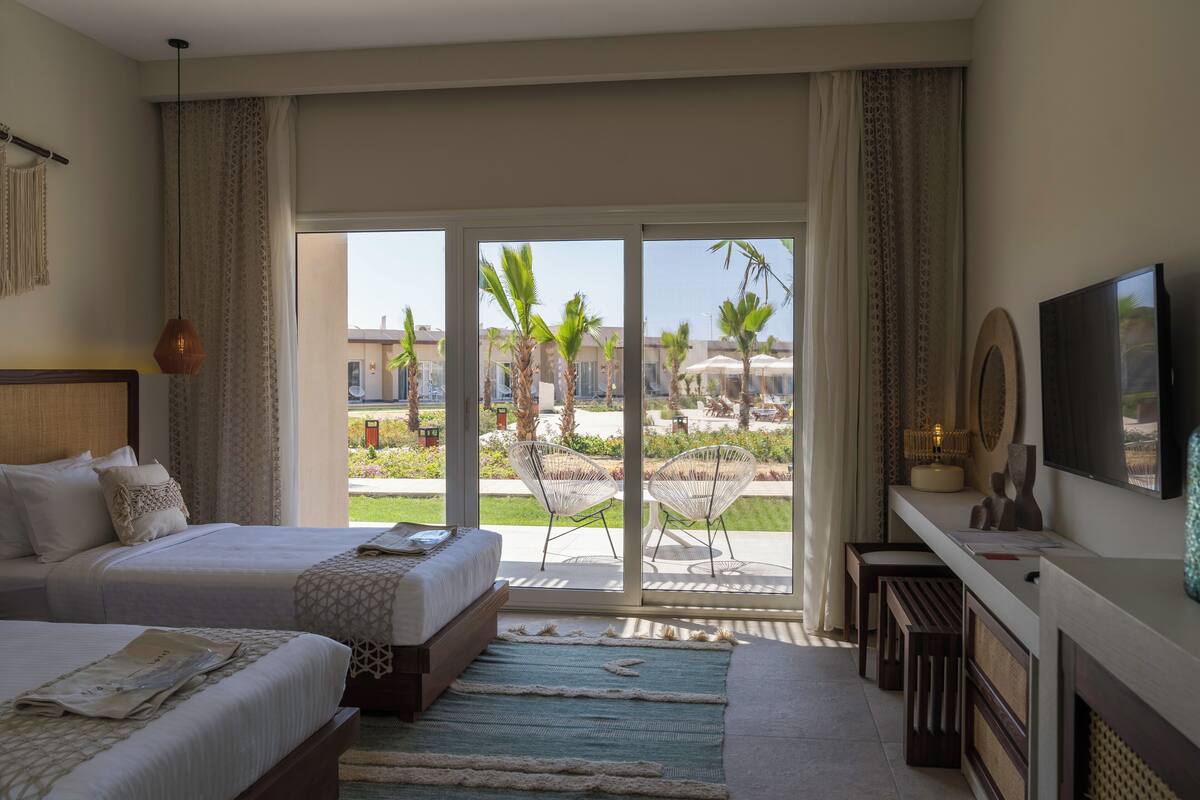 A hotel room with two beds, a large window with a view of palm trees and a pool, and a flat-screen TV on the wall.