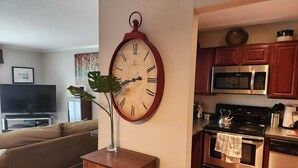A living room with a brown sofa, a wooden dresser, a large wall clock, a flat-screen TV, and a kitchen area with a microwave, stove, and oven. - Beautiful APT W Office Area 2BR Long Term (Elkins Park)