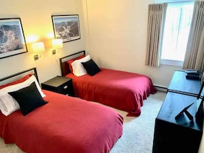 A hotel room with two beds, each with red bedding and black pillows, a small bedside table, a desk with a chair, and a window with curtains. - Mt Green Pool Gym Sauna Hot tub Open 3br 2b (Killington)