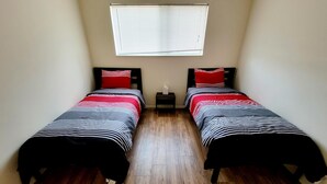 Een kleine, smalle kamer met twee bedden, elk voorzien van gestreept beddengoed en rode kussens. Er is een raam met zonwering, een klein tafeltje met een lamp en een houten vloer.