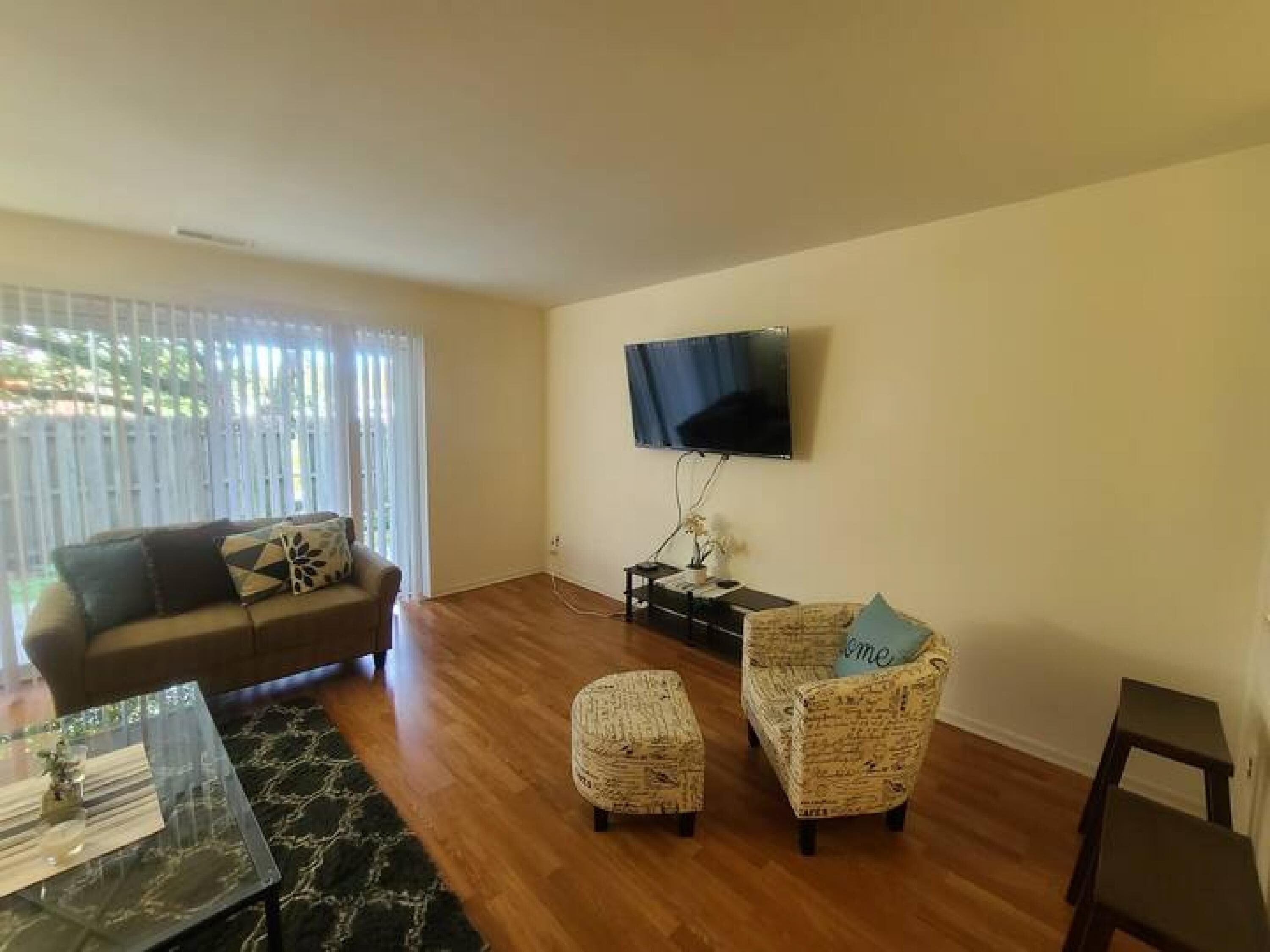 A living room with a sofa, ottoman, and armchair, a flat-screen TV mounted on the wall, and a dining table with chairs.