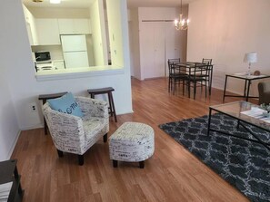 Uma sala de estar com sofá e apoio para os pés, uma mesa de jantar com cadeiras, cozinha ao fundo e um lustre.