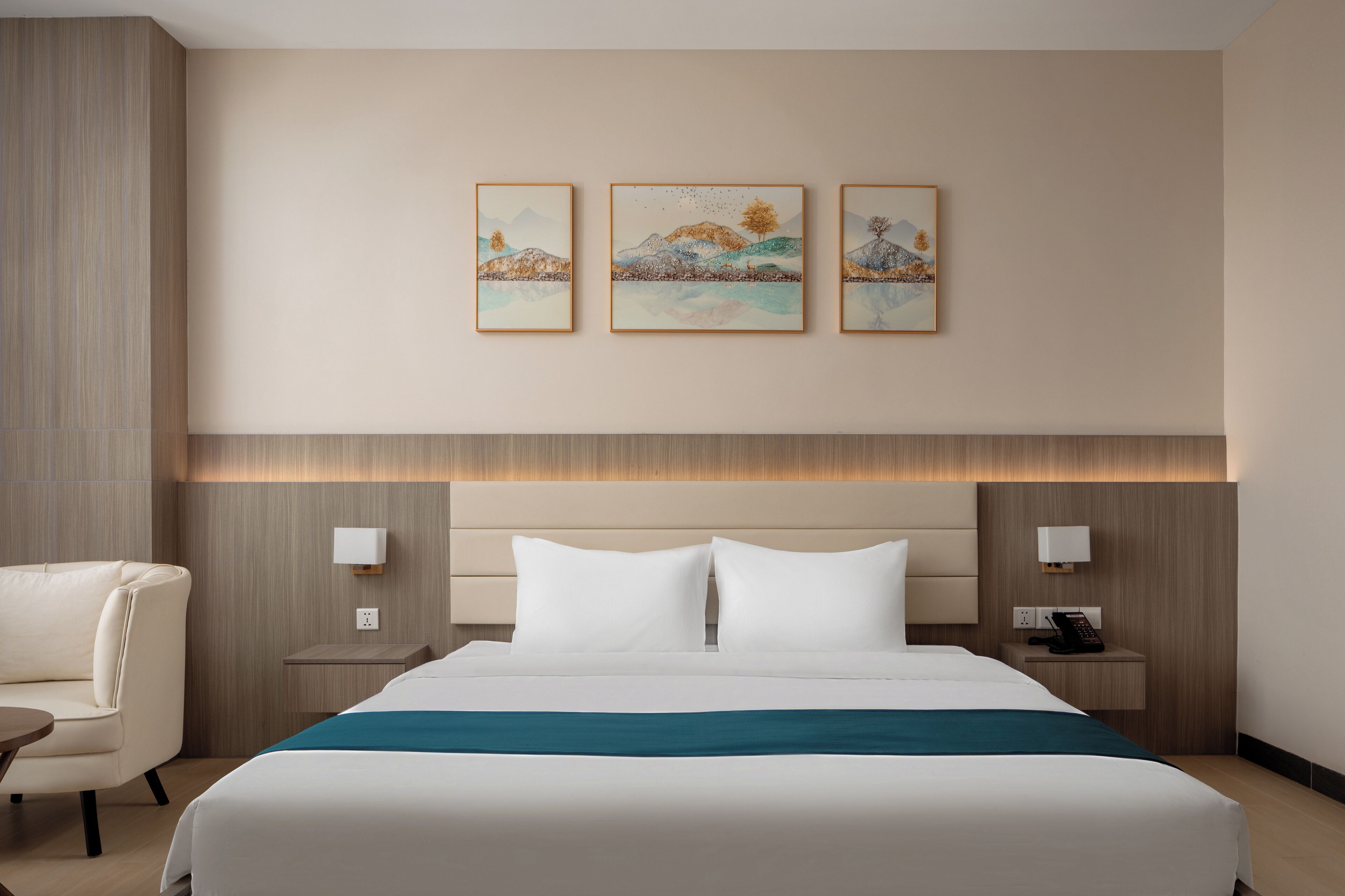 A hotel room with a bed, a chair, bedside tables, and three framed artworks on the wall.