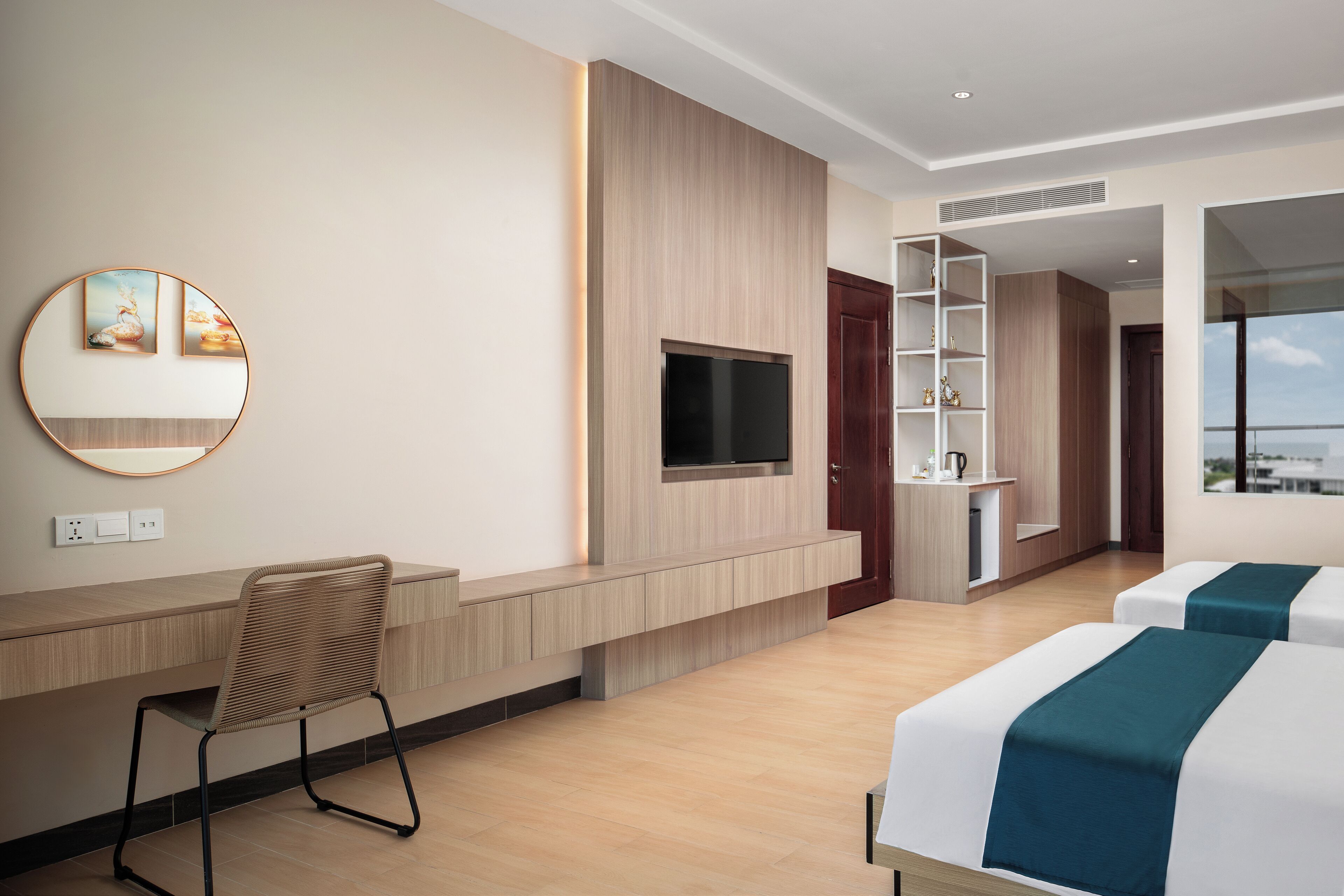 Una habitació d'hotel moderna amb una taula de treball de fusta, un televisor penjat a la paret, una zona de seients amb una cadira, dos llits amb llençols verds mares i una vista de la ciutat per la finestra.