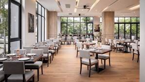 Een ruime restaurantruimte met meerdere eettafels en stoelen, grote ramen met uitzicht op groen, en plafondventilatoren.