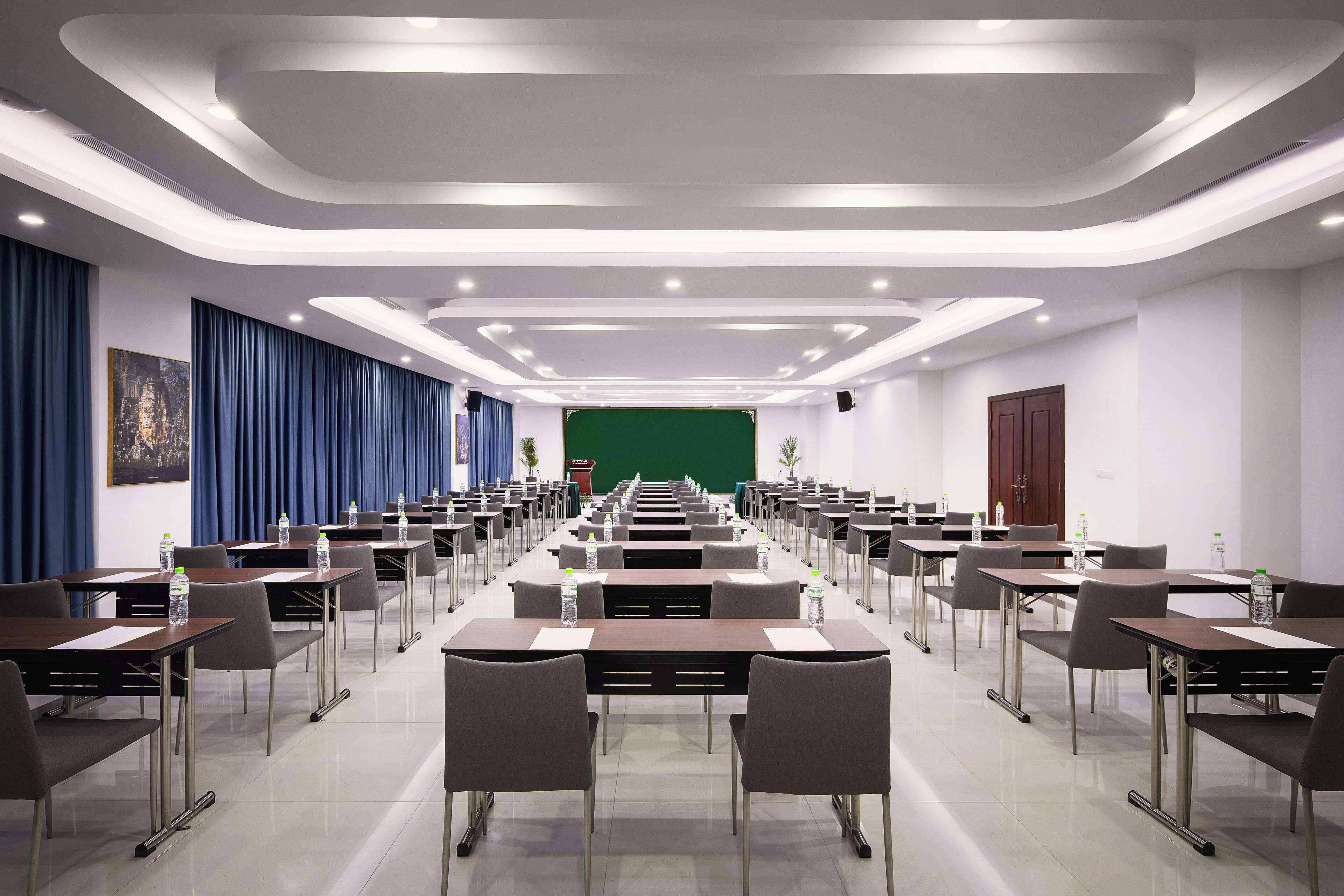 A conference room with rows of tables and chairs, a green screen at the front, and a modern ceiling design.