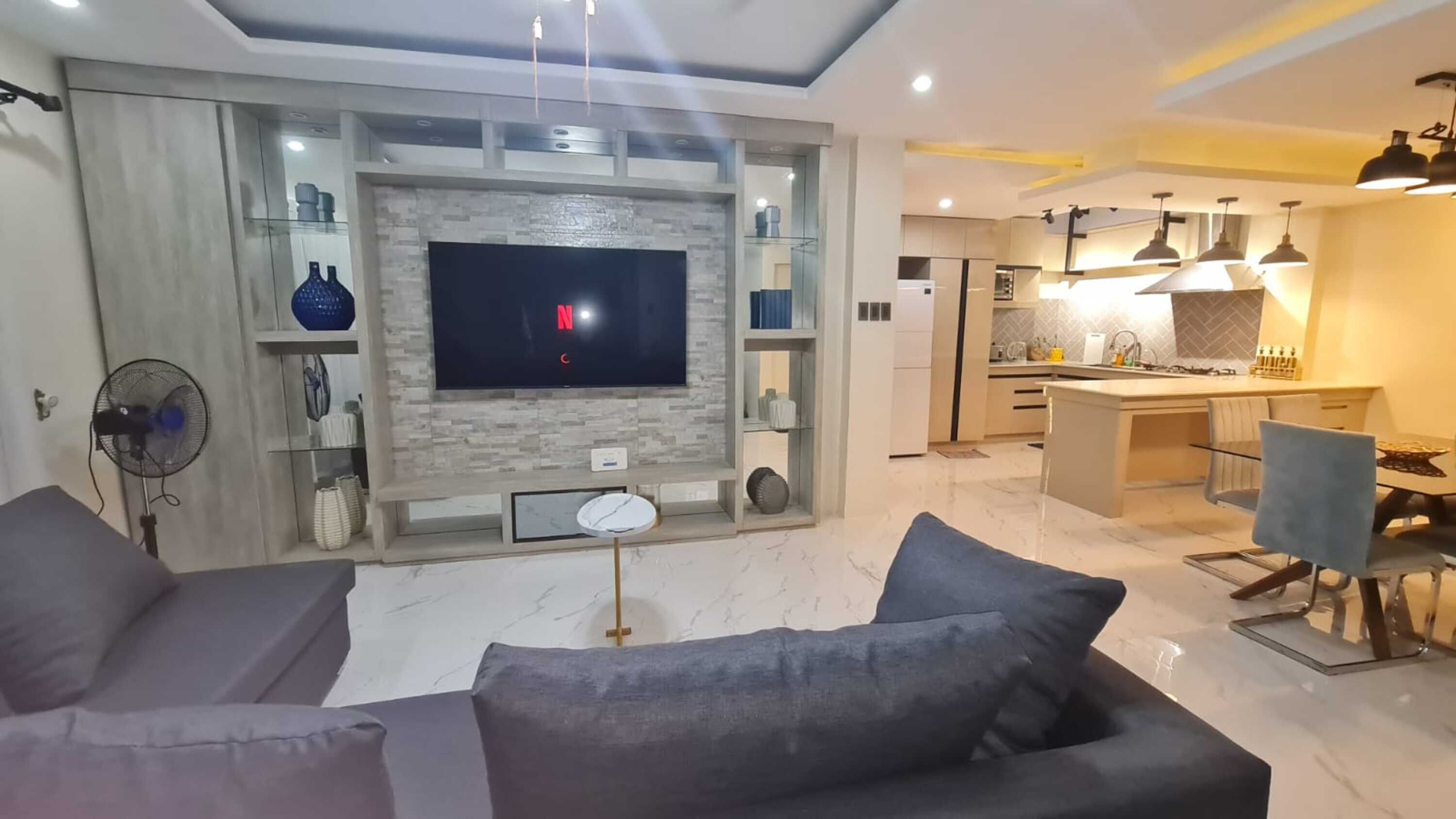 A modern living room with a flat-screen TV, grey sofas, and a dining area with a table and chairs.