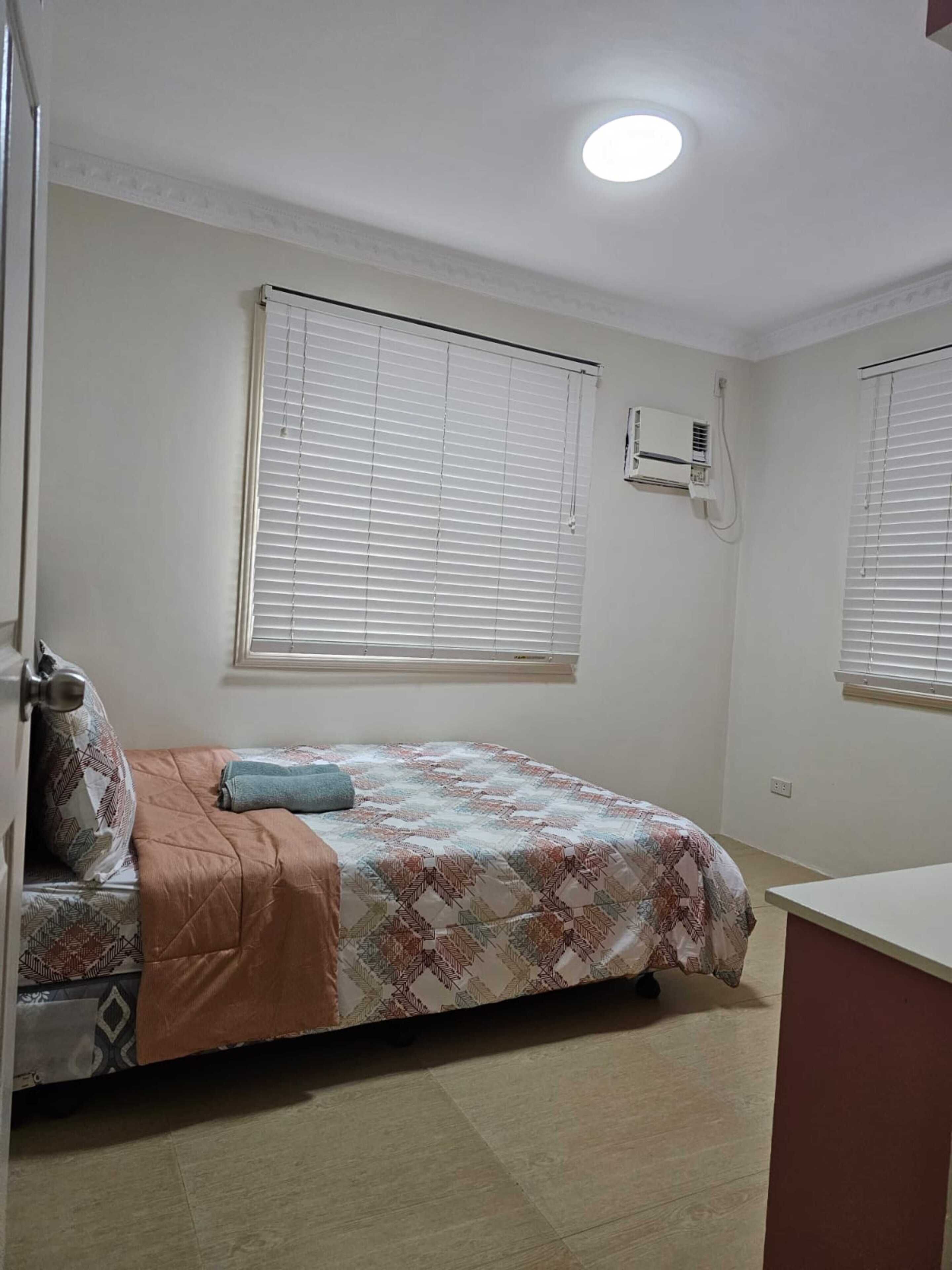 A bedroom with a bed, a window with blinds, an air conditioning unit, and a door.