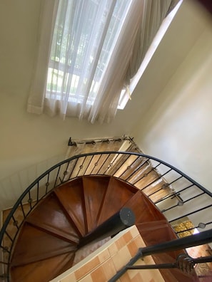 Uma escadaria em espiral com corrimão de madeira e balaústres de metal, conduzindo a um patamar com vista para o exterior.