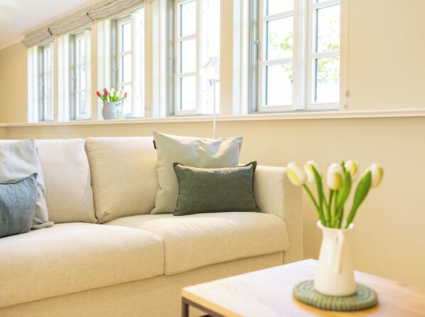 Ein Wohnzimmer mit einem beigen Sofa, einer Tulpenvase und einem Fenster mit Vorhängen.