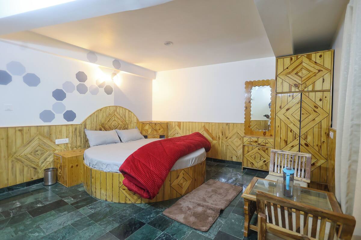 A bedroom with a wooden headboard, a bed with a red blanket, a small table with a kettle, and a mirror on the wall.