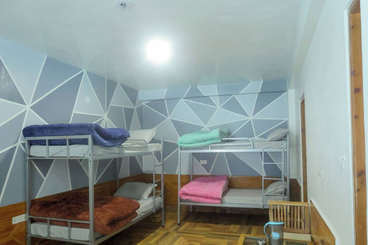 A dormitory room with bunk beds, a wooden floor, a small table with a chair, and a wall with geometric patterns.