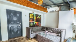Una habitación con un sofá seccional gris, una mesa de centro de vidrio, piso de madera, una puerta gris con detalles decorativos y una pared con un póster de Bob Marley.