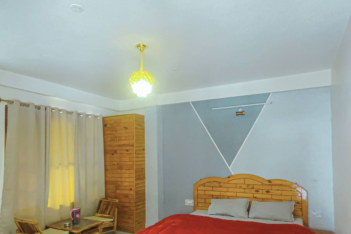 A bedroom with a wooden headboard, a red bedspread, a wooden nightstand, a wooden chair, a small table, and a curtain.