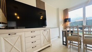 Un soggiorno moderno con una grande TV a schermo piatto, un mobiletto in legno con cassetti, un'area pranzo con tavolo e sedie e una vista attraverso ampie finestre.
