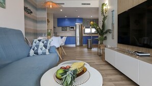 A modern living room with a blue sofa, a round coffee table with fruits, a TV, and a kitchen in the background. - CALA ALTA design apartment (Benidorm)