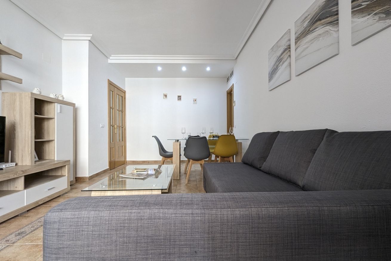 Ein modernes Wohnzimmer mit einem grauen Sofa, einem Glastisch und einer TV-Konsole. Im Hintergrund befindet sich ein Essbereich mit Tisch und Stühlen.