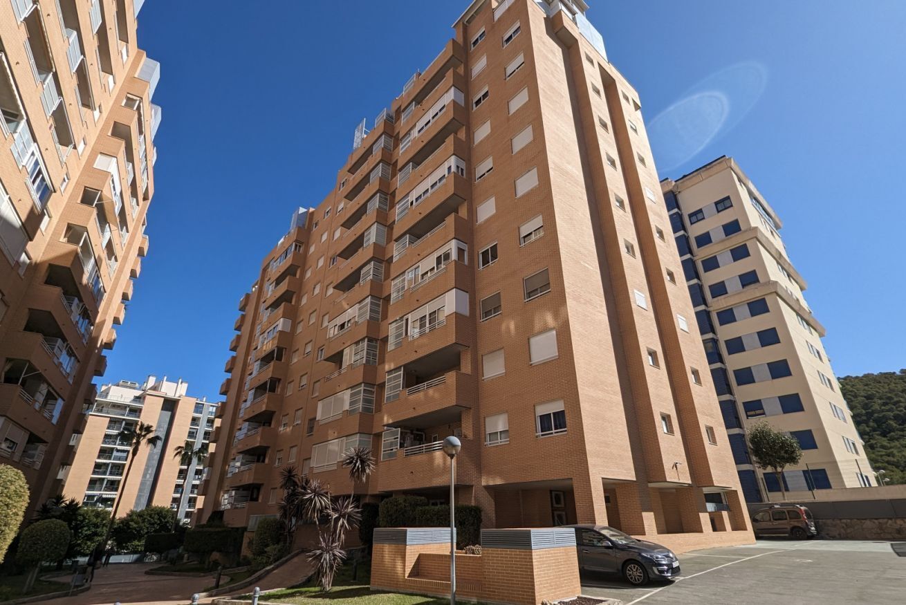 Un edificio residencial de varios pisos con balcones, un estacionamiento y cielo despejado.