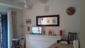Ein gemütlicher Wohnraum mit Deckenventilator, einem Fernseher auf einem weißen Schrank, einem kleinen Tisch mit Tasse und einem Sessel.