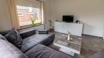 A modern living room with a grey sofa, a coffee table, a TV on a white cabinet, and a window with blinds.