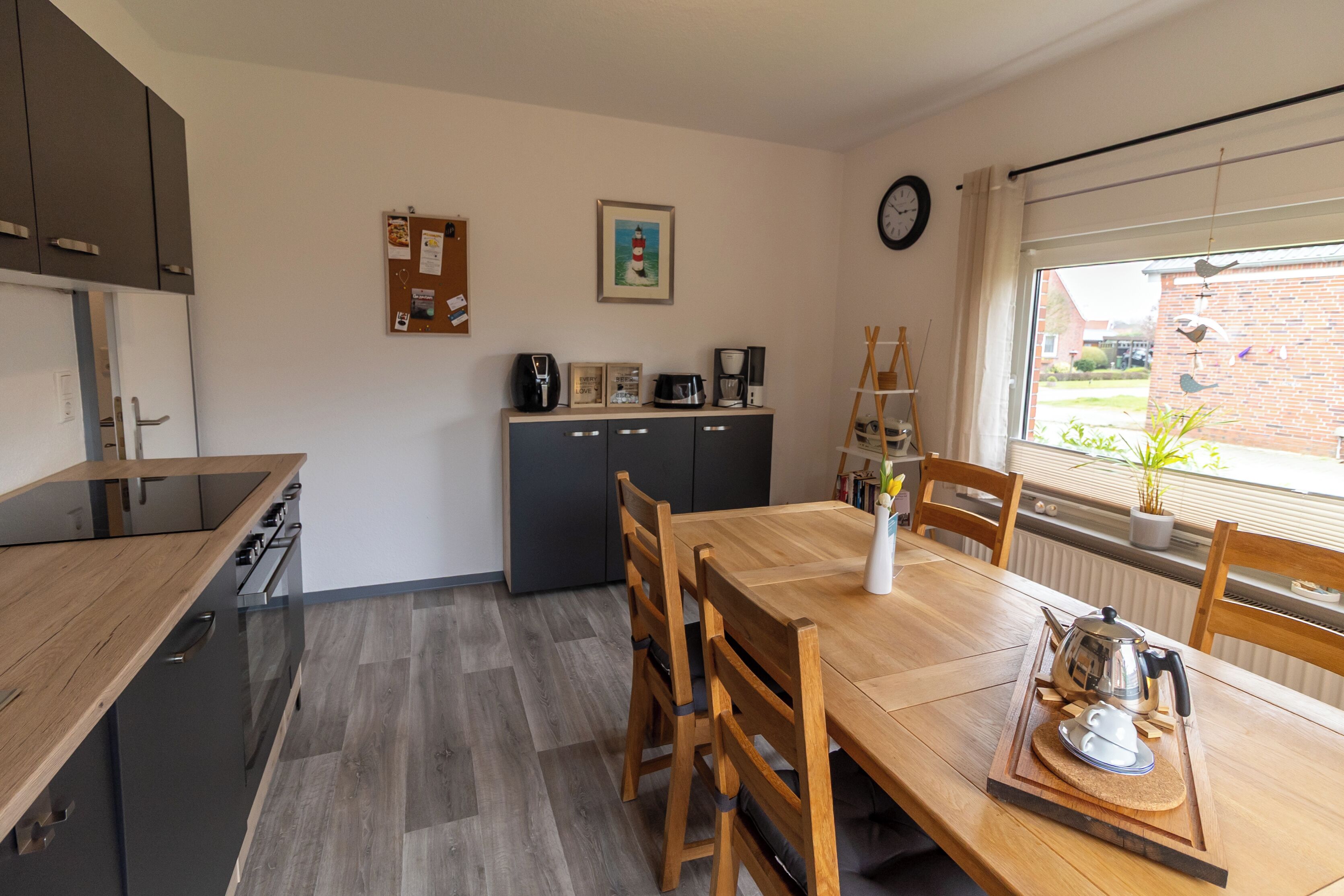 Une cuisine moderne avec une table à manger en bois, des meubles de rangement sombres et une fenêtre donnant sur un bâtiment en briques.