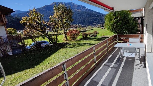 A balcony with a table and chairs, a view of a mountain, and a parked car. - Gemütliches Appartement für Erholung und Sport (Klosters-Serneus)