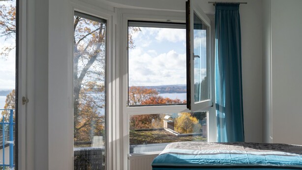 A bedroom with a large window, a bed with patterned bedding, and a view of trees and a body of water.
