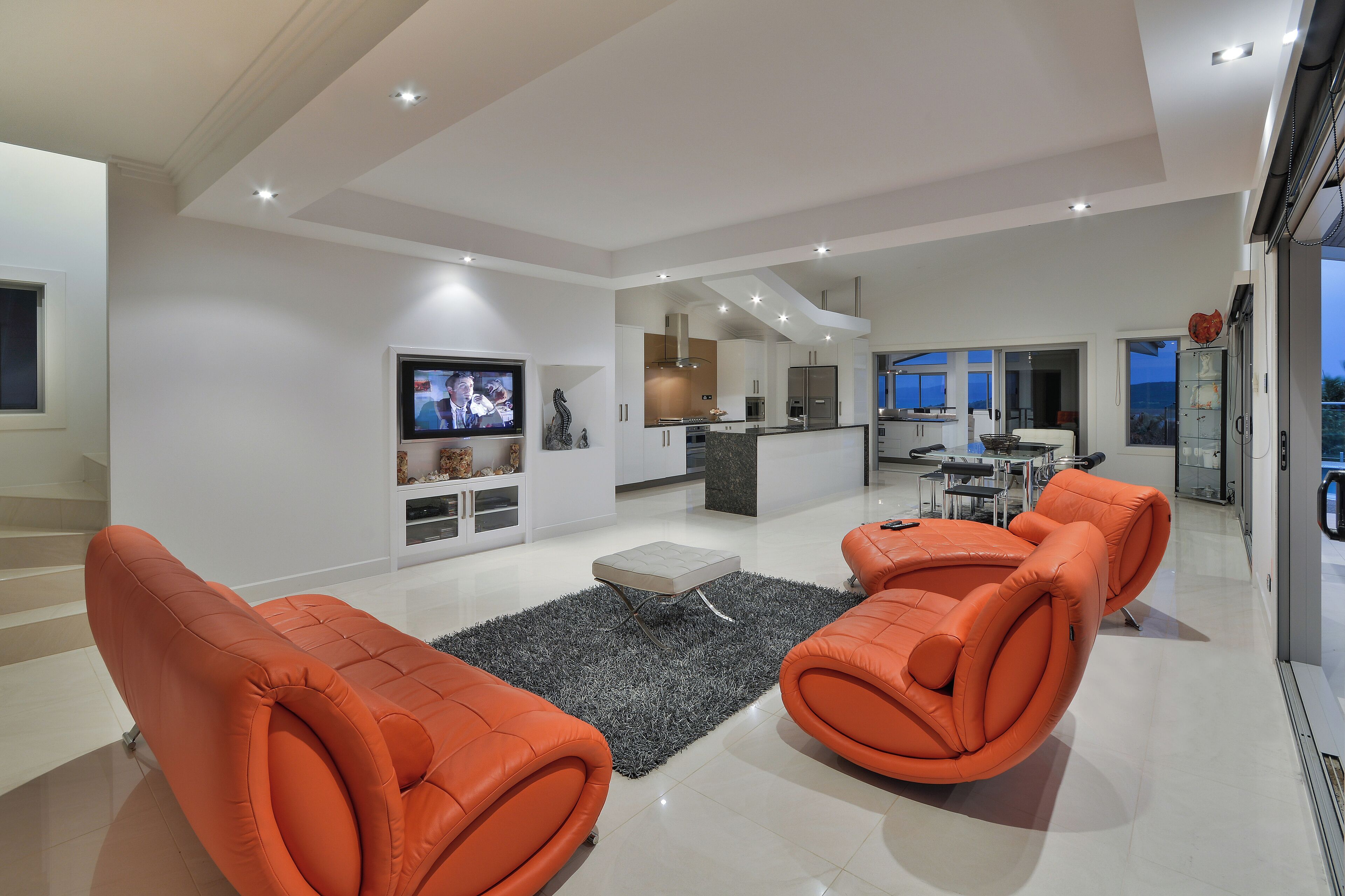 A modern living room with orange sofas, a flat-screen TV, and a kitchen in the background.