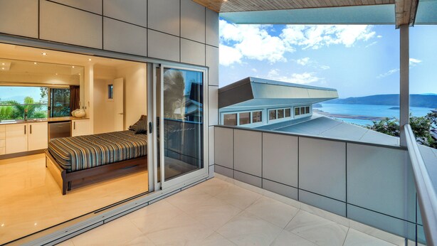 A modern bedroom with a large bed, sliding glass doors leading to a balcony with a view of a distant building and landscape. - Jasmine house  (Hamilton Island)