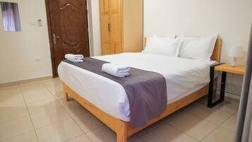 Una cama bien hecha con una manta gris y almohadas blancas, una mesita de noche de madera con una lámpara, y una puerta de madera al fondo.