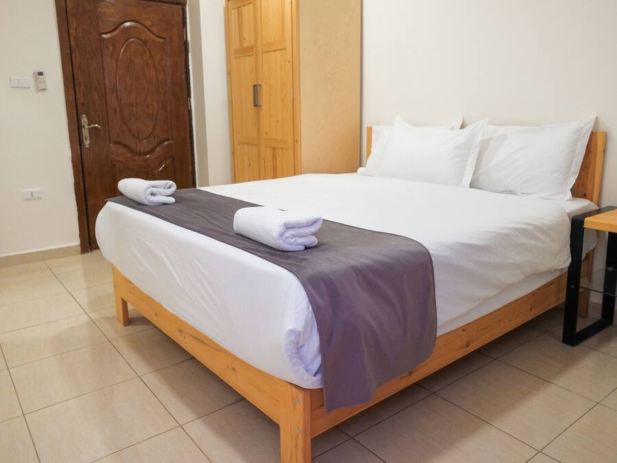 Una cama bien hecha con una manta gris y almohadas blancas, una mesita de noche de madera con una lámpara, y una puerta de madera al fondo.