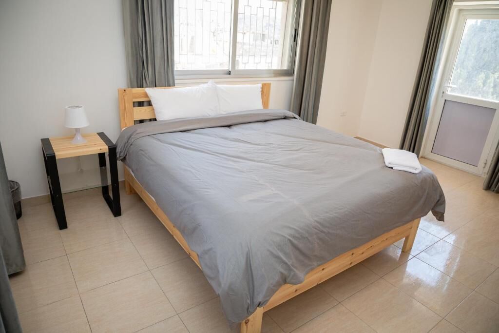 A neatly made bed with a grey blanket and white pillow in a room with a window, a bedside table with a lamp, and a tiled floor.