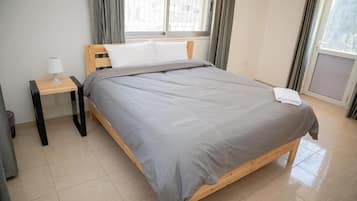 A neatly made bed with a grey blanket and white pillow in a room with a window, a bedside table with a lamp, and a tiled floor.