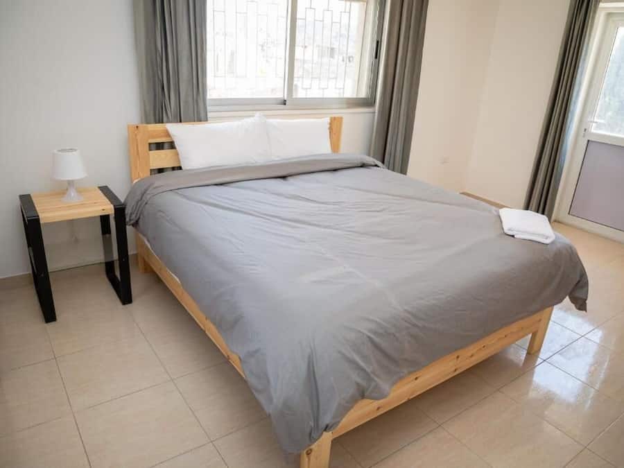 A neatly made bed with a grey blanket and white pillow in a room with a window, a bedside table with a lamp, and a tiled floor.