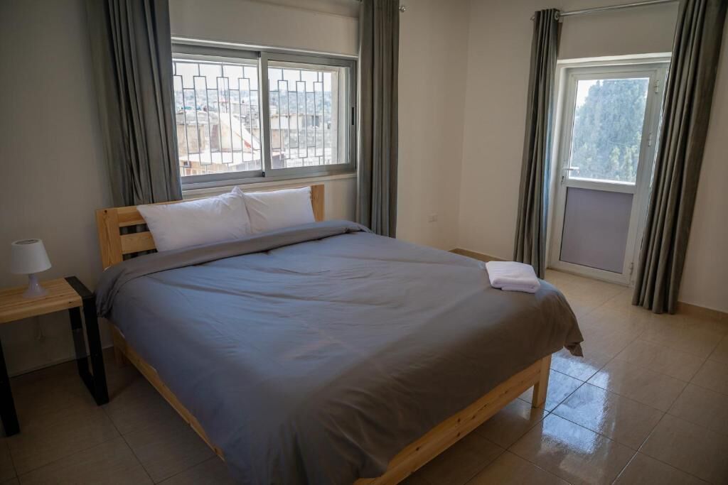 Una cama bien hecha con cubrecama gris y almohadas blancas, una mesita de noche con lámpara y una ventana con cortinas.