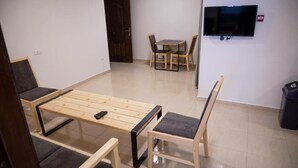 A room with a wooden bed frame, two chairs, a small table, a TV, and a door. - Dali House (Amman)